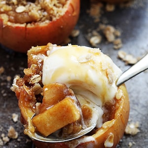 May include: A close-up of a baked apple with a scoop of vanilla ice cream on top. The apple is filled with a cinnamon crumble topping and drizzled with caramel sauce.