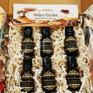 May include: A box filled with six small bottles of essential oil. The bottles are labeled with different scents, including Cinnamon Bun, Lemon Pound Cake, Snickerdoodle, and Blueberry Cheesecake. The box is lined with brown paper and has a small card on top that says "Bakers Kitchen Premium Home Fragrance Oils".