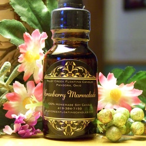 May include: A small, dark brown glass bottle with a gold label that reads "Riley Creek Floating Candles, Pandora, Ohio, Cranberry Marmalade, 100% Homemade Soy Candle, 419-384-7150, RileyCreekFloatingCandles.com". The bottle is surrounded by pink and green flowers.