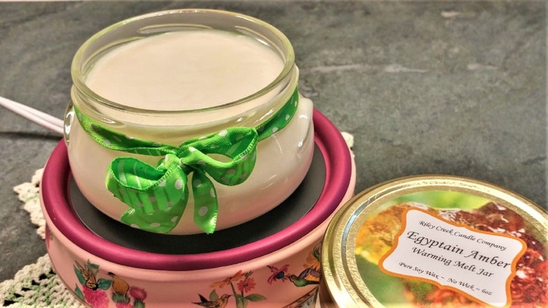 May include: A glass jar of wax melt with a green polka dot ribbon tied around the top. The jar is sitting on a pink and white warming dish. The jar is labelled "Egyptian Amber Warming Melt Jar" with the text "Riley Creek Candle Company" above the label.