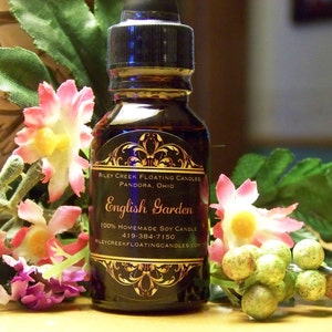 May include: A small dark brown glass bottle with a gold label that reads "Riley Creek Floating Candles Pandora, Ohio English Garden 100% Homemade Soy Candle 419-384-7150 rileycreekfloatingcandles.com". The bottle is sitting on a wooden surface with pink and purple flowers.