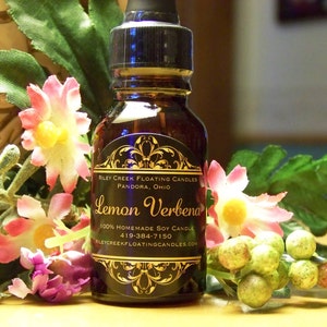 May include: A small glass bottle with a black label that reads "RILEY CREEK FLOATING CANDLES PANDORA, OHIO Lemon Verbena 100% HOMEMADE SOY CANDLE 419-384-7150 BLEYCREEKFLOATINGCANDLES.COM". The bottle is surrounded by green leaves and pink flowers.