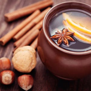 May include: A brown ceramic mug filled with mulled wine, garnished with orange slices and a star anise. Cinnamon sticks and hazelnuts are scattered around the mug.