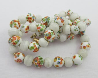 Puede incluir: Un collar de cuentas de vidrio blanco con estampado floral y cierre dorado. Las cuentas alternan entre blanco y blanco con un estampado floral.