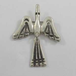May include: A silver bird brooch with outstretched wings and a dangling tail. The bird's wings and tail feature detailed etched patterns. The brooch has a pin clasp on the back for secure attachment to clothing or accessories.