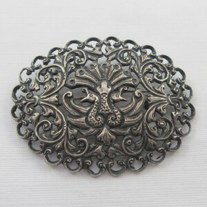 May include: An oval-shaped silver brooch with a detailed design of two peacocks with their tails spread out. The brooch is made of filigree work and has a delicate, intricate design.