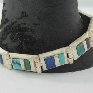 Può includere: Bracciale color argento con intarsi rettangolari di pietre turchesi, blu e verdi. Il bracciale è esposto su una superficie nera, evidenziando il suo design geometrico e i colori vivaci. Chiusura a cerniera.