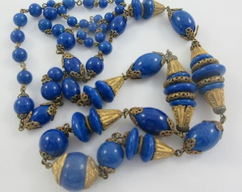 May include: A long blue and gold beaded necklace with a variety of bead sizes and shapes. The necklace features a mix of round, oval, and cylindrical beads, all with a deep blue color. The beads are interspersed with gold spacers and accents, adding a touch of elegance to the design.