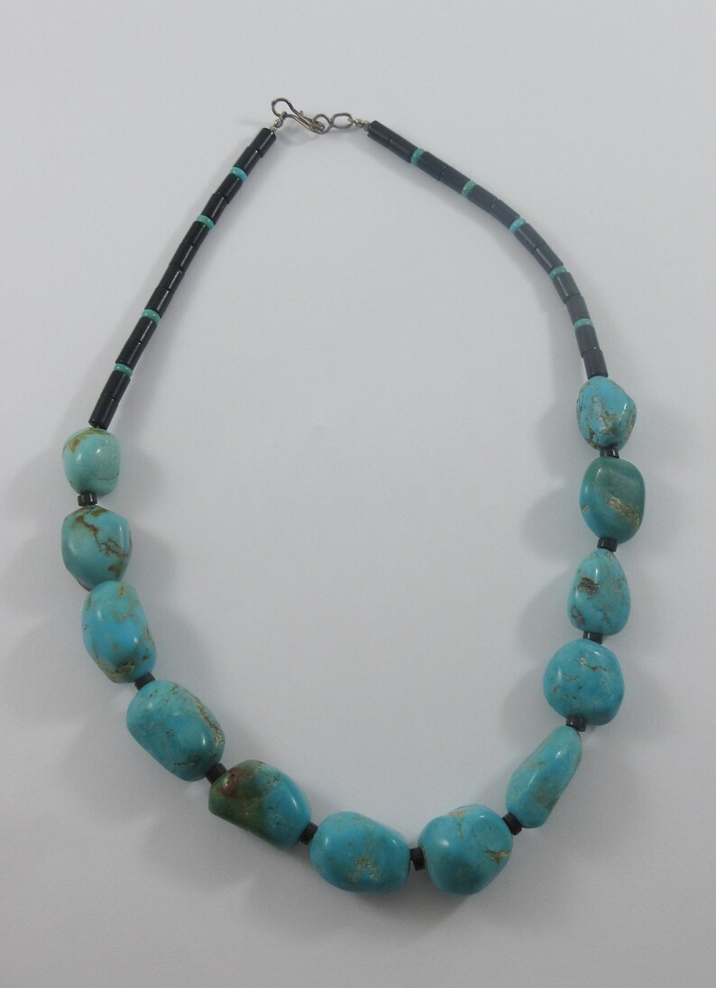 May include: A turquoise and black beaded necklace with a silver clasp. The necklace is made of large, irregular turquoise stones separated by small black beads.