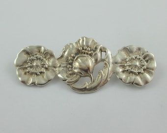 May include: A silver brooch with three stylized poppy flowers. The center flower is larger and has a more detailed design.