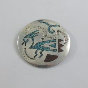 Puede incluir: Colgante redondo de plata con incrustaciones de turquesa y marrón. El colgante presenta una figura estilizada de ave o humana, formas geométricas y líneas decorativas. La incrustación de turquesa contrasta con la plata.