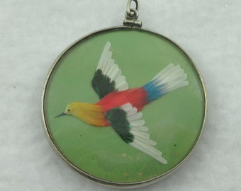 Op de afbeelding: Een zilveren ovale hanger met een groene achtergrond en een kleurrijke vogel gemaakt van veren. De vogel heeft gele, rode, blauwe, groene en witte veren.