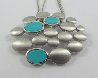 Op de afbeelding: Een zilveren ketting met een hanger gemaakt van meerdere met elkaar verbonden ovale vormen. De hanger heeft twee turquoise stenen.