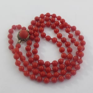 May include: A long strand of round, vibrant red beads strung together with knots between each bead. The necklace features a decorative, circular gold-toned metal clasp that is set with a single, matching round red bead at its center.