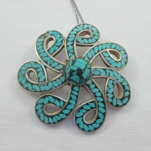 Pu&ograve; includere: Una collana con un ciondolo in argento con un designo a forma di fiore turchese e nero. Il fiore &egrave; fatto di intricati vortici e ha una pietra turchese centrale.