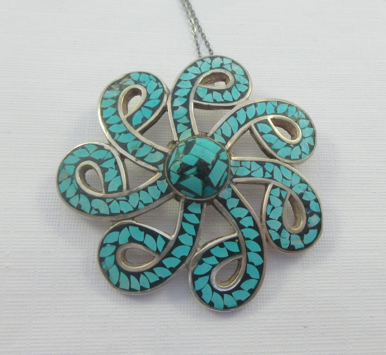 Pu&ograve; includere: Una collana con un ciondolo in argento con un designo a forma di fiore turchese e nero. Il fiore &egrave; fatto di intricati vortici e ha una pietra turchese centrale.