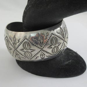 May include: A wide, silver cuff bracelet with intricate geometric patterns. The bracelet features a series of diamond shapes, each containing detailed designs. The metal has a polished finish, reflecting light. The bracelet is displayed on a black surface.