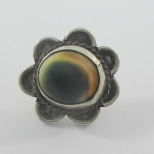 May include: A silver-toned ring with a flower-shaped setting. The ring features a central oval stone with a dark green and tan color pattern. The setting has a rope-like border and petal-shaped accents.