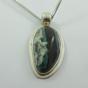 Puede incluir: Un colgante de plata con una piedra de ágata verde, negra y blanca. La piedra está engastada en un bisel de plata y tiene un acabado liso y pulido.