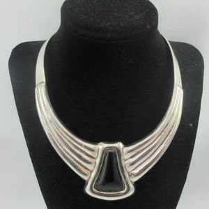 Op de afbeelding: Een zilveren choker ketting met een zwarte steen in het midden. De ketting heeft een geometrisch ontwerp en is gemaakt van meerdere lagen zilver.