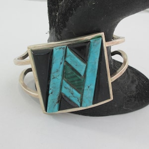 May include: A silver cuff bracelet featuring a square centerpiece with geometric inlay. The design consists of black onyx, bright blue turquoise, and green malachite segments arranged in a diagonal pattern. The metal band is open and polished.