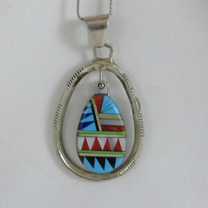 May include: A silver pendant necklace featuring a colorful, inlaid stone pendant. The pendant has a teardrop shape with a mosaic design in shades of blue, red, white, and black. The pendant is suspended within a silver oval frame.