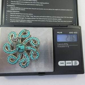 Pu&ograve; includere: Un ciondolo in turchese e argento a forma di polpo con una catena, che pesa 2,16 grammi su una bilancia digitale. La bilancia indica "AWS-600" e ha una capacit&agrave; di 600 grammi.