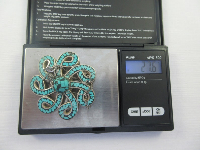 Pu&ograve; includere: Un ciondolo in turchese e argento a forma di polpo con una catena, che pesa 2,16 grammi su una bilancia digitale. La bilancia indica "AWS-600" e ha una capacit&agrave; di 600 grammi.