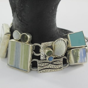Könnte beinhalten: Ein silbernes Armband mit rechteckigen und quadratischen Anhängern. Die Anhänger zeigen eine Vielzahl von Farben, darunter Grün, Blau und Weiß, und einige haben Muschel- und Steinakzente. Das Armband wird auf einer schwarzen Form präsentiert.