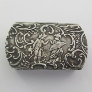 Pode incluir: Uma caixa retangular de metal prateado com uma cena em relevo de duas figuras em trajes históricos, uma delas segurando um leque, rodeadas por intrincados ornamentos de volutas e motivos florais em toda a superfície superior da tampa.