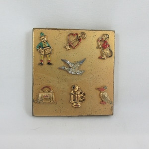May include: A gold-colored metal compact with a variety of colorful enamel designs, including a bird, a monkey, a heart with a key, a purse, a lamp, and a pelican.