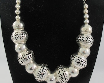 May include: Silver-toned necklace with a series of large, round beads interspersed with intricately patterned beads. The necklace is displayed on a black velvet bust, showcasing its design and craftsmanship. The chain is made of small, round beads.