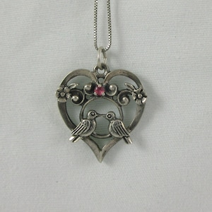 May include: Silver-toned pendant necklace featuring a heart-shaped design with two kissing birds. The pendant has floral accents and a small red gemstone. The necklace hangs on a delicate chain, perfect for everyday wear.