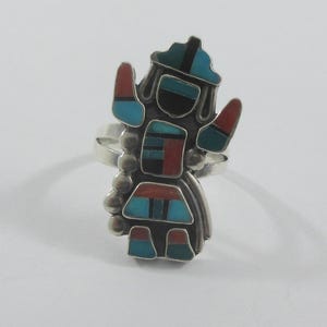 May include: A silver ring featuring a Native American Kachina doll design. The ring has a detailed figure with inlaid turquoise, red, black, and green stones. The ring's design includes a hat, arms, and a skirt-like shape.