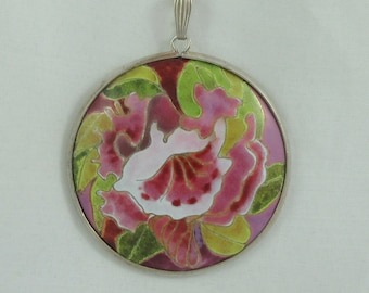 Op de afbeelding: Een zilveren ketting met een ronde hanger met een roze en groen bloemmotief. De hanger is gemaakt van cloisonné-email.