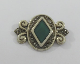 May include: Vintage silver-tone brooch with a central green diamond-shaped stone. The oval frame is embellished with small, sparkling accents and decorative scrollwork on each side, creating an elegant accessory.