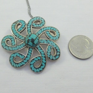 Pu&ograve; includere: Una collana con un ciondolo in argento con un designo a forma di fiore in turchese e argento. Il fiore &egrave; fatto di intricati vortici e ha un centro turchese.