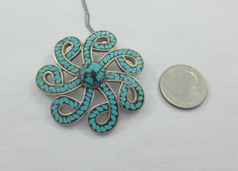 Pu&ograve; includere: Una collana con un ciondolo in argento con un designo a forma di fiore in turchese e argento. Il fiore &egrave; fatto di intricati vortici e ha un centro turchese.