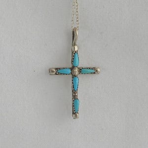 May include: A silver cross pendant necklace with turquoise teardrop-shaped stones. The cross has a simple design with a small chain. The turquoise stones are set in the arms of the cross and in the center.