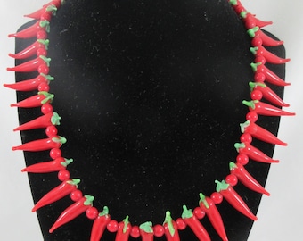 May include: A vibrant red chili pepper necklace displayed on a black mannequin. The necklace features glossy red chili pepper pendants with green accents, strung together with small red beads. A unique and eye-catching accessory.