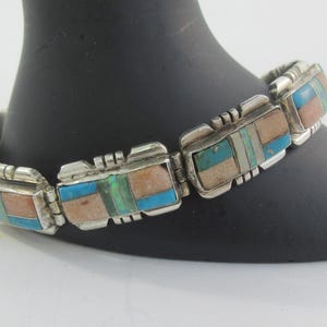 May include: A link bracelet crafted from silver metal, featuring rectangular segments inlaid with rectangular pieces of blue turquoise, tan stone, and iridescent green stone. The metal links are connected by hinges, creating a flexible band.