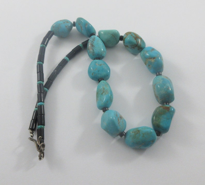 May include: A turquoise and black beaded necklace with a silver clasp. The necklace is made of large, irregular turquoise stones and small, black beads.