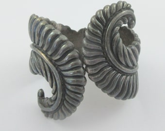 Puede incluir: Un brazalete de plata con un diseño de hoja en espiral. El brazalete está hecho de dos piezas que están conectadas por una bisagra.