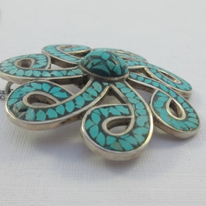 Pu&ograve; includere: Una collana d'argento con un ciondolo a forma di fiore turchese. Il fiore &egrave; fatto di intricati anelli d'argento ed &egrave; intarsiato con pietre turchesi.