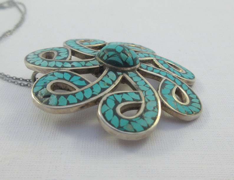 Pu&ograve; includere: Una collana d'argento con un ciondolo a forma di fiore turchese. Il fiore &egrave; fatto di intricati anelli d'argento ed &egrave; intarsiato con pietre turchesi.
