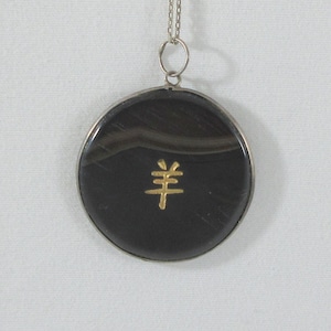 May include: A round pendant necklace with a silver chain. The pendant features a black stone with a gold-colored Chinese character. The stone is encased in a silver bezel.