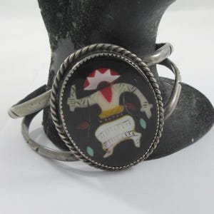 May include: Silver cuff bracelet with a central oval design. The design features a figure with red, white, and yellow accents on a black background, framed by a twisted silver rope. The bracelet has a simple, elegant design.