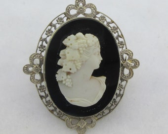 May include: A white cameo brooch featuring a woman's profile with a floral crown, set in a silver filigree frame with a black enamel background.