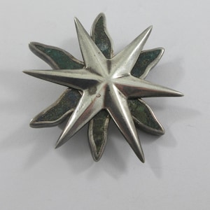 May include: A silver-coloured star-shaped brooch with eight points. The brooch has a central star design with a textured surface. The base of the star has a green-coloured, leaf-like design.