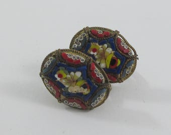 Puede incluir: Un par de pendientes de mosaico ovalados. Los pendientes presentan un diseño floral con azulejos rojos, amarillos, blancos y azules. El mosaico está engastado en un marco de metal dorado con un borde decorativo.
